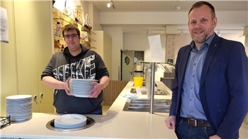 Zwei Männer stehen an einer Theke. Der linke hält einen Stapel Teller in seinen Händen. 