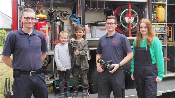 Zwei Jungen von 6 und 4 Jahren stehen zusammen mit zwei Feuerwehrmännern und einem jugendlichen Mitglied der Jugendwehr vor einem Löschfahrzeug.   