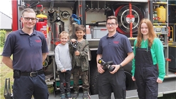 Zwei Jungen von 6 und 4 Jahren stehen zusammen mit zwei Feuerwehrmännern und einem jugendlichen Mitglied der Jugendwehr vor einem Löschfahrzeug.   