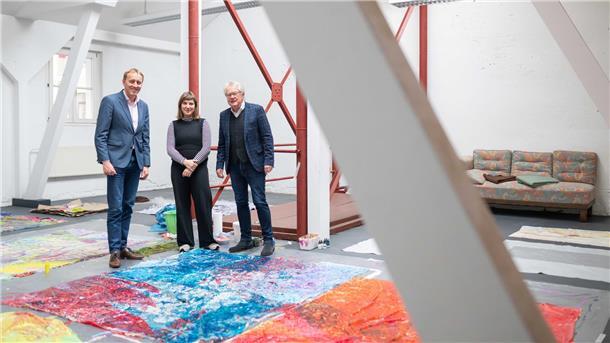 Drei Personen stehen in einem großzügigen, hellen Atelierraum zwischen großformatigen, farbenfrohen Kunstwerken, die auf dem Boden ausgebreitet sind. Im Hintergrund sind rote Stahlträger, weiße Wände und ein Sofa zu sehen.