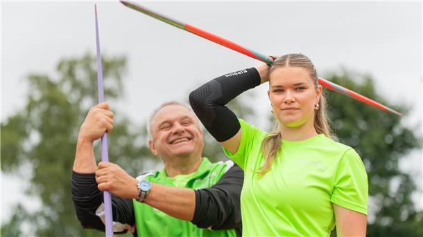 Ein Trainer hält einen Speer, während sein Schützling einen Speer über den Kopf hält.