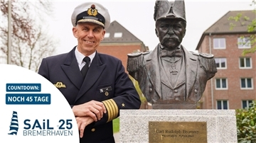 Ein Marineoffizier in Uniform steht lächelnd neben einer Büste von Carl Rudolph Brommy; im Vordergrund ein Countdown zur Sail 2025 in Bremerhaven mit „Noch 45 Tage“.