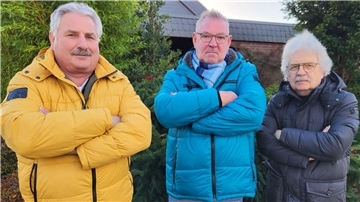 Drei Männer stehen in Winterjacken mit verschränkten Armen vor einem Wohngebiet. 
