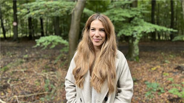 Eine Frau mit langen, braunen Haaren und einem beigefarbenen Mantel steht in einem Wald.