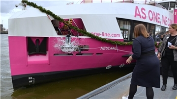Eine Frau lässt eine Sektflasche am Bug einer pink-weiß gestrichenen Fähre mit dem Namen "Neuland" zerschellen.