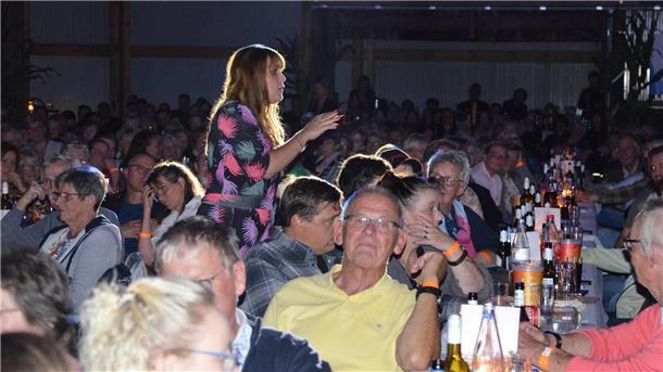 Eine Frau im bunten Kleid steht mitten im Publikum und spricht, während zahlreiche Menschen an langen Tischen sitzen, Getränke vor sich haben und aufmerksam zuhören.