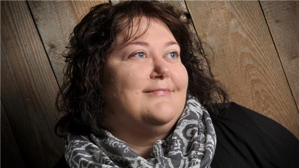 Eine Frau mit Halstuch guckt zur Seite und lächelt leicht. Sie steht vor einer mit Holz verkleideten Wand.
