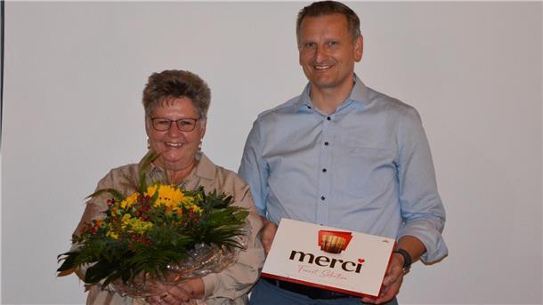 Veronika Ratje wird mit einem Blumenstrauß und einer Schachtel Pralinen verabschiedet. Neben ihr steht ein Mann, der die Dankesgabe überreicht. Beide lächeln.