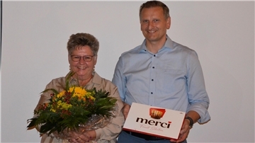 Veronika Ratje wird mit einem Blumenstrauß und einer Schachtel Pralinen verabschiedet. Neben ihr steht ein Mann, der die Dankesgabe überreicht. Beide lächeln.