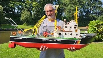 Ein Mann mit grauen Haaren hält ein schwarz-rotes Modellschiff in den Händen. 