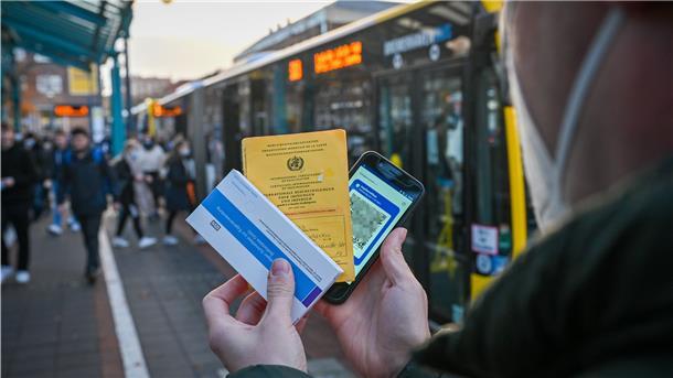 Ein Mann hält einen Corona-Schnelltest, seinen Impfausweis und ein Handy in der Hand.