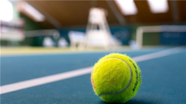 Ein gelber Tennisball liegt auf blauem Teppichboden in einer Tennishalle.
