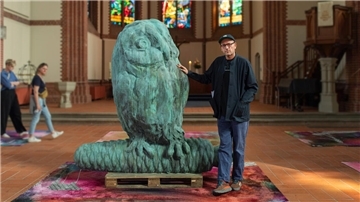 In einer Kirche steht eine mehr als zwei Meter große Eule aus Bronze, daneben steht ein Mann. Auf dem Boden liegen bunte Teppiche.