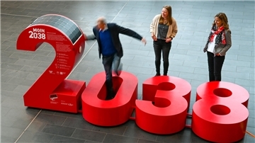 Drei Personen, ein Mann und zwei Frauen, interagieren mit einer großen, roten Installation in Form der Zahl "2038"