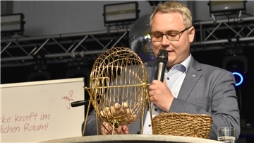 Ein Mann hat einen runden Korb aus Metall vor sich auf einem Tisch stehen und spricht in ein Mikrofon.