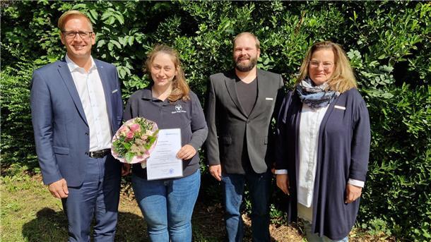 Vier Menschen stehen vor einer grünen Hecke. Landrat Stephan Siefken beglückwünscht Wiebke Mayer-Reinecke zu ihrer neuen Aufgabe als Bezirksschornsteinfegermeisterin.