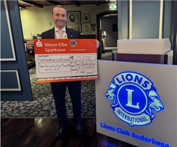 Ein Mann im Anzug hält einen großen symbolischen Spendenscheck über 25.000 Euro in den Händen. Neben ihm steht ein beleuchtetes Schild mit dem Logo des Lions Club Bederkesa.