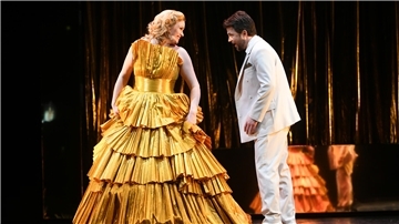 Szenenbild aus "Die lustige Witwe": Eine Frau in goldenem Kleid und ein Mann in weißem Anzug stehen auf der Bühne und singen. 