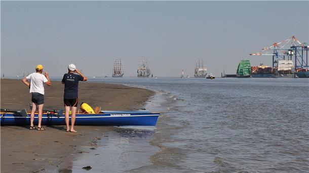 Bild von Ruderern mit ihrem Boot auf einer Sandbank in der Weser. Im Hintergrund sind Großsegler und die Hafenkräne in Bremerhaven zu sehen.