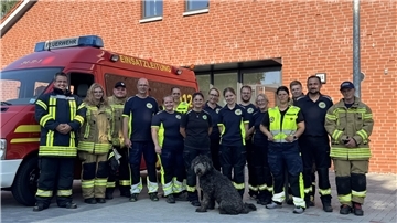 Einsatzkräfte stehen vor einem Gebäude. Ein Hund sitzt davor. 