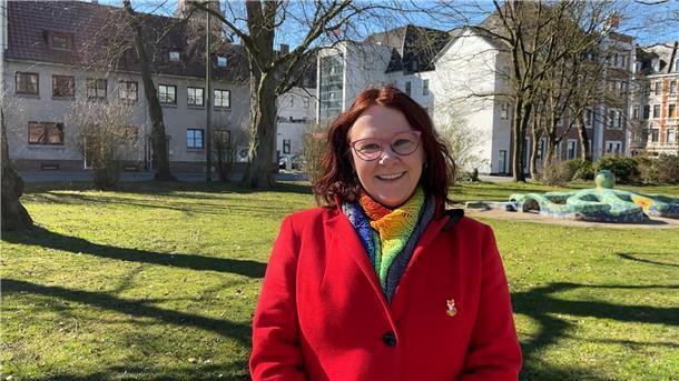 Eine Frau in einem roten Mantel steht in einem Park und lächelt in die Kamera. 