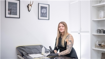 Eine junge tätowierte Frau mit langen blonden Haaren hält eine kabellose Tätowiermaschine in der Hand. 