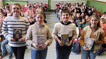 Vier Kinder stehen mit jeweils einem Buch in der Hand vor Schülerinnen und Schülern. 
