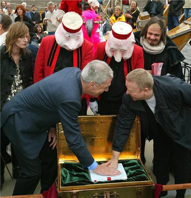 Zwei Männer, die in eine goldene Truhe fassen. Hinter ihnen stehen Personen, die Masken auf dem Kopf tragen.