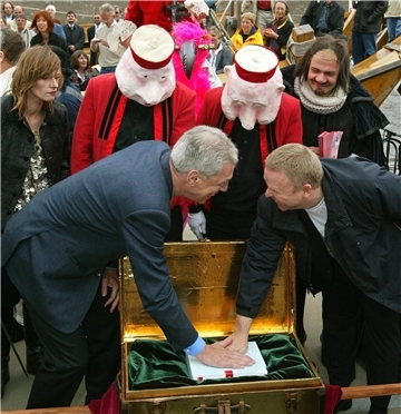 Zwei Männer, die in eine goldene Truhe fassen. Hinter ihnen stehen Personen, die Masken auf dem Kopf tragen.
