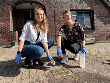 Zwei junge Frauen hocken am Boden. Vor ihnen sechs kleine Messingplatten – die Stolpersteine.