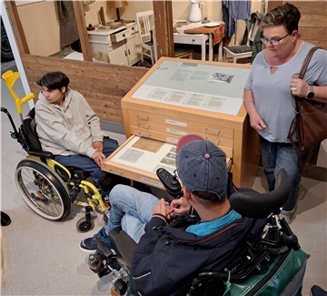 Zwei Menschen im Rollstuhl und eine Frau betrachten in einem Museum eine Schubladen-Vitrine mit historischen Objekten. Die Szene zeigt einen inklusiven Museumsbesuch.
