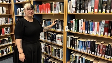 Eine Frau steht in der Gemeindebücherei Loxstedt vor einem Bücherregal und greift zu einem Buch. Sie lächelt und trägt ein schwarzes Kleid sowie Brille und Piercings.