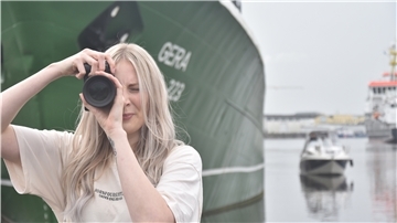 Eine Frau mit Kamera steht vor einem Schiff und einem Boot und fotografiert.
