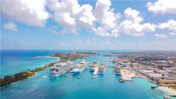 Luftaufnahme des Kreuzfahrthafens von Nassau auf den Bahamas: Mehrere große Kreuzfahrtschiffe liegen nebeneinander an den Piers. Im Hintergrund erstreckt sich die Stadt mit Hotels und Gebäuden, links das türkisfarbene Meer und Sandstrände.