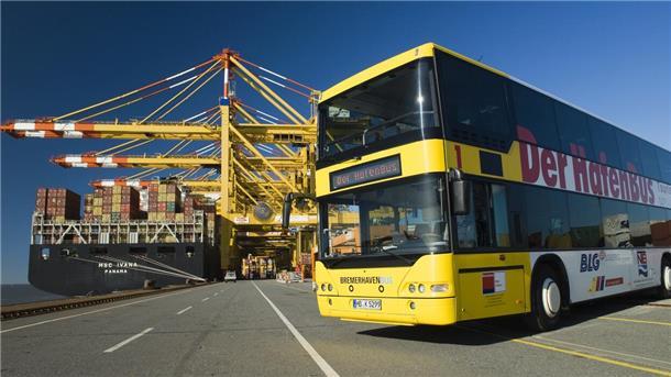 Ein gelber Doppeldecker Bus steht vor einem Containerschiff im Hafen. Die Sonne scheint.