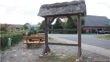 Die alte, leere Ortstafel in Wittstedt steht ohne Beschilderung an der Ecke von Kreisstraße und Ortsstraße. Das Holzgestell mit Reetdach ist sichtbar gealtert, im Hintergrund sind eine Sitzbank, Straßen und Bauernhäuser zu sehen.