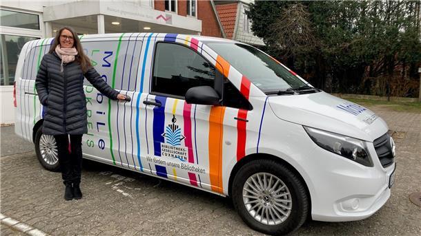 Eine Frau mit langer, dunkler Jacke und Schal steht neben einem bunt gestalteten Fahrzeug, das die Aufschrift „MobiLe Landkreis“ trägt.