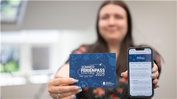 Eine Frau hält eine Karte mit der Aufschrift „Sommerferienpass 2025“ sowie ein Smartphone mit geöffneter Website des Ferienpass-Portals in die Kamera.