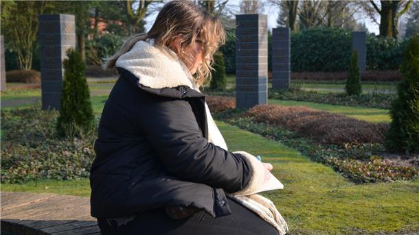 Eine junge Frau sitzt auf einem Friedhof und schreibt einen Text auf einen Block. 