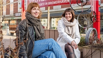 Links sitzt eine Frau mit roten Haaren, Lederjacke und einem karierten Schal, rechts sitzt eine Frau mit dunklen Haaren, einem grauen Mantel und beigefarbenen Schal. Beide sitzen in einem hölzernen Pavillon vor dem Stadttheater in Bremerhaven.