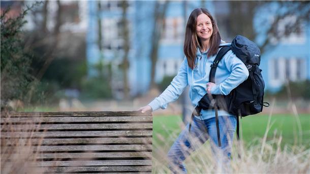 Eine Frau mit braunen Haaren und einer blauen Jacke steht mit einem Wanderrucksack im Grünen: Wanderliebhaberin Maria Luisa Kuzmic aus Bremerhaven.