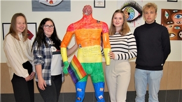 Eine in Regenbogenfarben bemalte Figur ohne Kopf steht in der Mitte. Links und rechts davon stehen drei Schülerinnen und ein Schüler der Waldschule Hagen-Beverstedt.