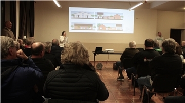 Bürgerinnen und Bürger sitzen in einem Versammlungsraum und verfolgen eine Präsentation zu Entwürfen des neuen Feuerwehrhauses in Kassebruch, die per Beamer an die Wand projiziert werden.