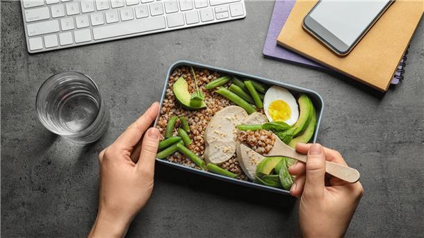 Meal Prep für Berufstätige: Gesund essen und Zeit sparen ist keine Zauberei Eine Meal Prep Box mit Bohnen, Ei, Getreide, Avocado und Tofu stehen auf einem grauen Tisch, auf dem auch ein Glas Wasser, eine weiße Tastatur und Hefte liegen.