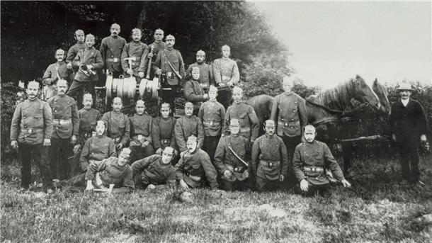 Schwarz-weiß-Foto einer historischen Feuerwehrgruppe aus dem frühen 20. Jahrhundert. Etwa 25 Männer in einheitlicher Uniform mit Pickelhauben posieren vor einem von Pferden gezogenen Löschwagen mit Handdruckspritze. Im Hintergrund Bäume und Gebüsch, im Vordergrund sitzen einige Feuerwehrmänner auf dem Gras.