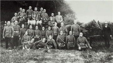 Schwarz-weiß-Foto einer historischen Feuerwehrgruppe aus dem frühen 20. Jahrhundert. Etwa 25 Männer in einheitlicher Uniform mit Pickelhauben posieren vor einem von Pferden gezogenen Löschwagen mit Handdruckspritze. Im Hintergrund Bäume und Gebüsch, im Vordergrund sitzen einige Feuerwehrmänner auf dem Gras.