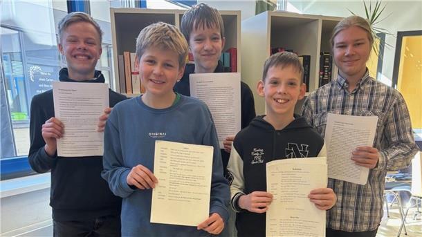 Fünf Jungen stehen lächelnd nebeneinander und halten Zettel in den Händen. 