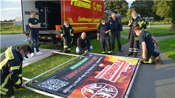 Mehrere Feuerwehrmänner und Feuerwehrfrauen stehen um das Werbe-Banner herum.