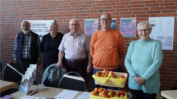 Fünf ältere Frauen und Männer stehen lächelnd nebeneinander vor einer Backsteinwand. Vor ihnen stehen Tische mit Informationsmaterial, Äpfeln in Körben und Flyern.