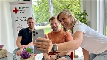 Eine Frau hält ein Handy und lacht, zwei Männer schauen auf das Display.